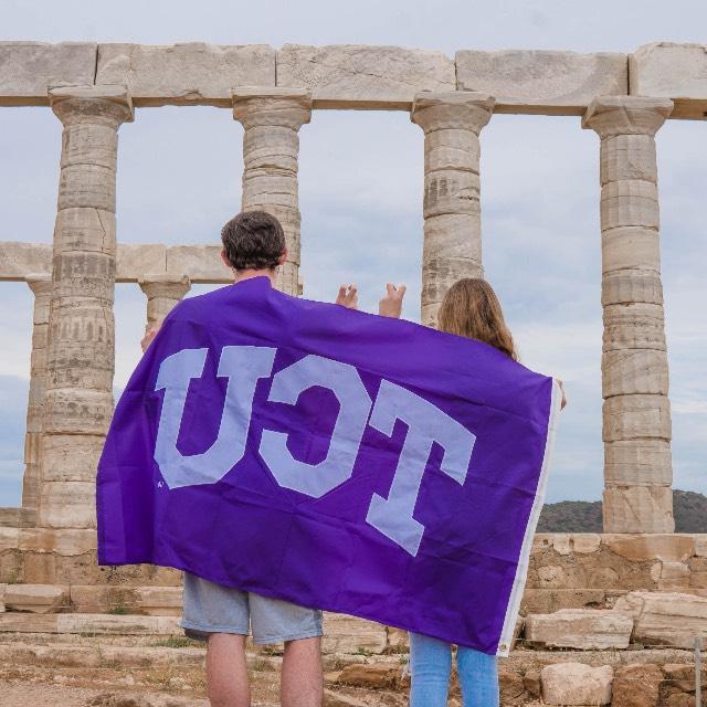 两个学生, 背对着我们, 当他们看着一座古典纪念碑的柱子时，将TCU的旗帜挂在肩上